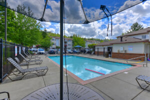 Exterior Swimming Pool, photo taken form bistro table, chaise tanning chairs around pool, fenced in, meticulous landscaping alongside fence, trees and residential buildings in background, photo taken on a sunny day.
