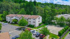 Aerial exterior, residential building, three-story walk-up, basketball court, dense woods behind property, parking out front.